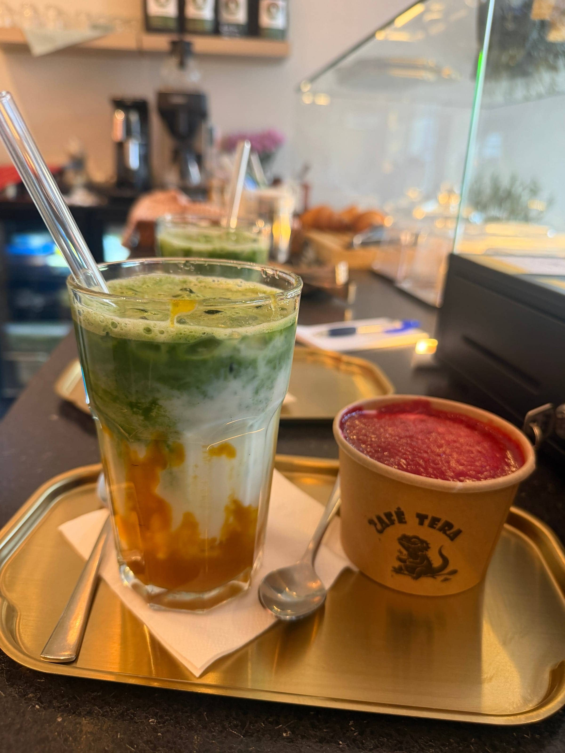 Iced Mango Matcha im Glas und Himbeer-Tiramisu im Café-Tera-Becher auf einem Messingtablett an der Theke