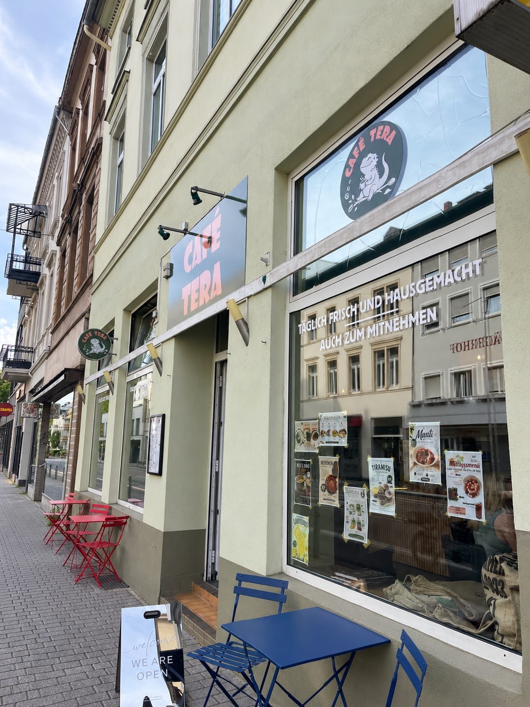 Ladenfront von Café Tera in der Bahnhofstraße mit Logo-Schildern und dem Schaufenster-Schriftzug „Täglich frisch und hausgemacht – auch zum Mitnehmen“, davor Bistrotische auf dem Gehweg