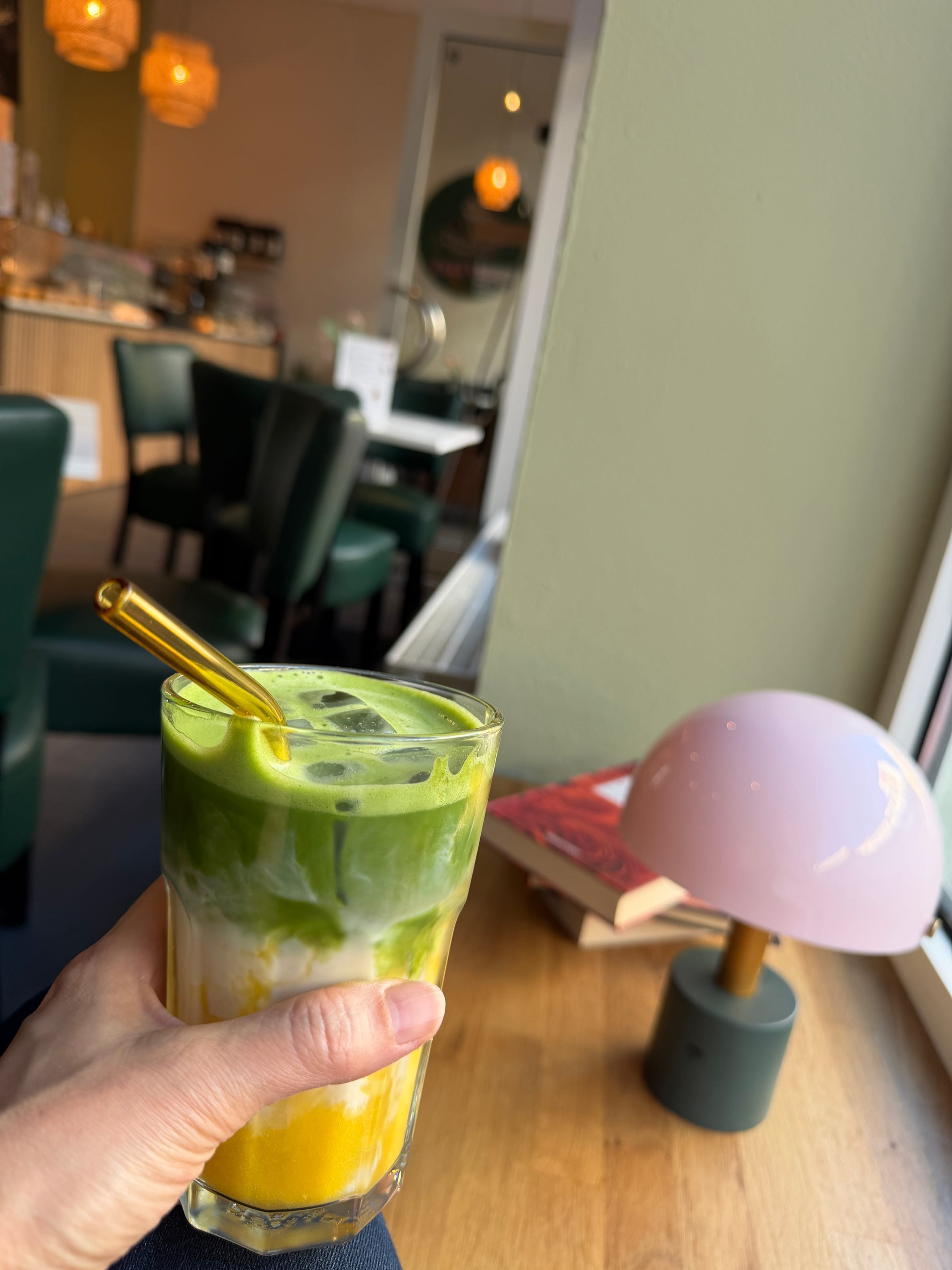 Iced Mango Matcha in der Hand am Fensterplatz neben einer rosa Tischleuchte, im Hintergrund dunkelgrüne Stühle, Rattanleuchten und die Theke