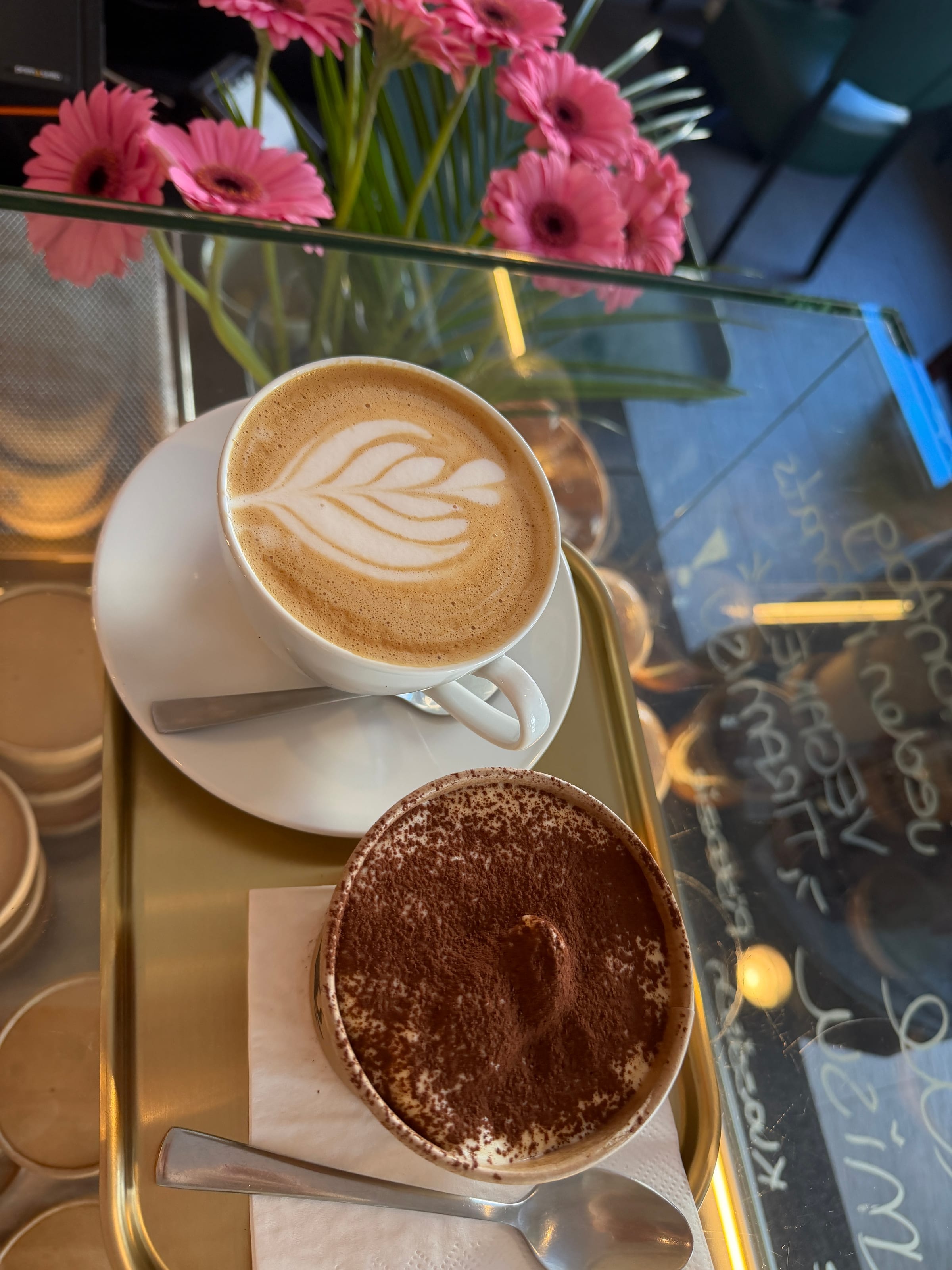 Kaffee mit Latte Art und klassisches Tiramisu mit Kakao auf einem Messingtablett, dahinter rosa Gerbera auf der Vitrine