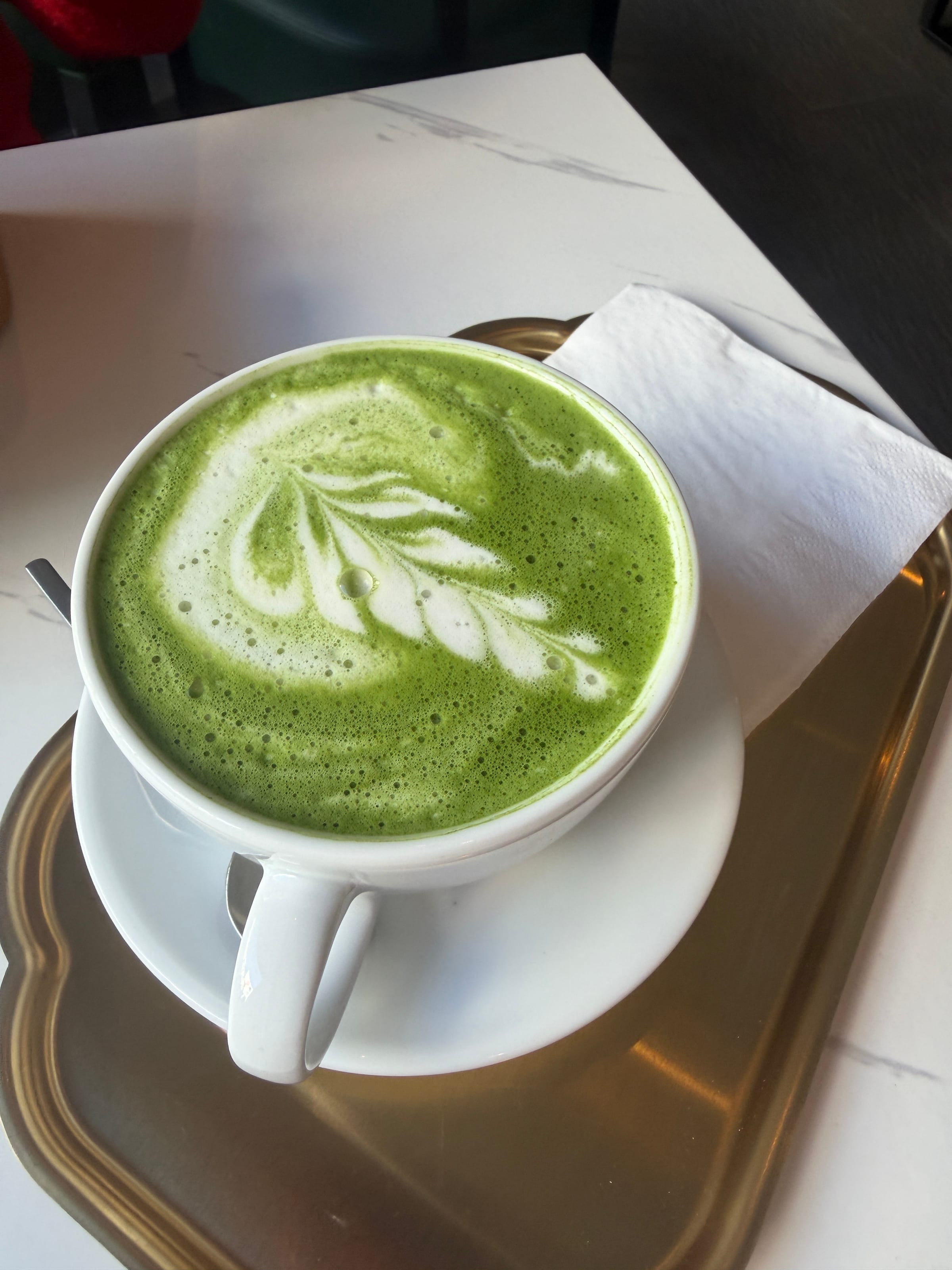 Matcha Latte mit Latte Art in einer weißen Tasse auf einem Messingtablett, daneben eine Serviette auf dem Marmortisch