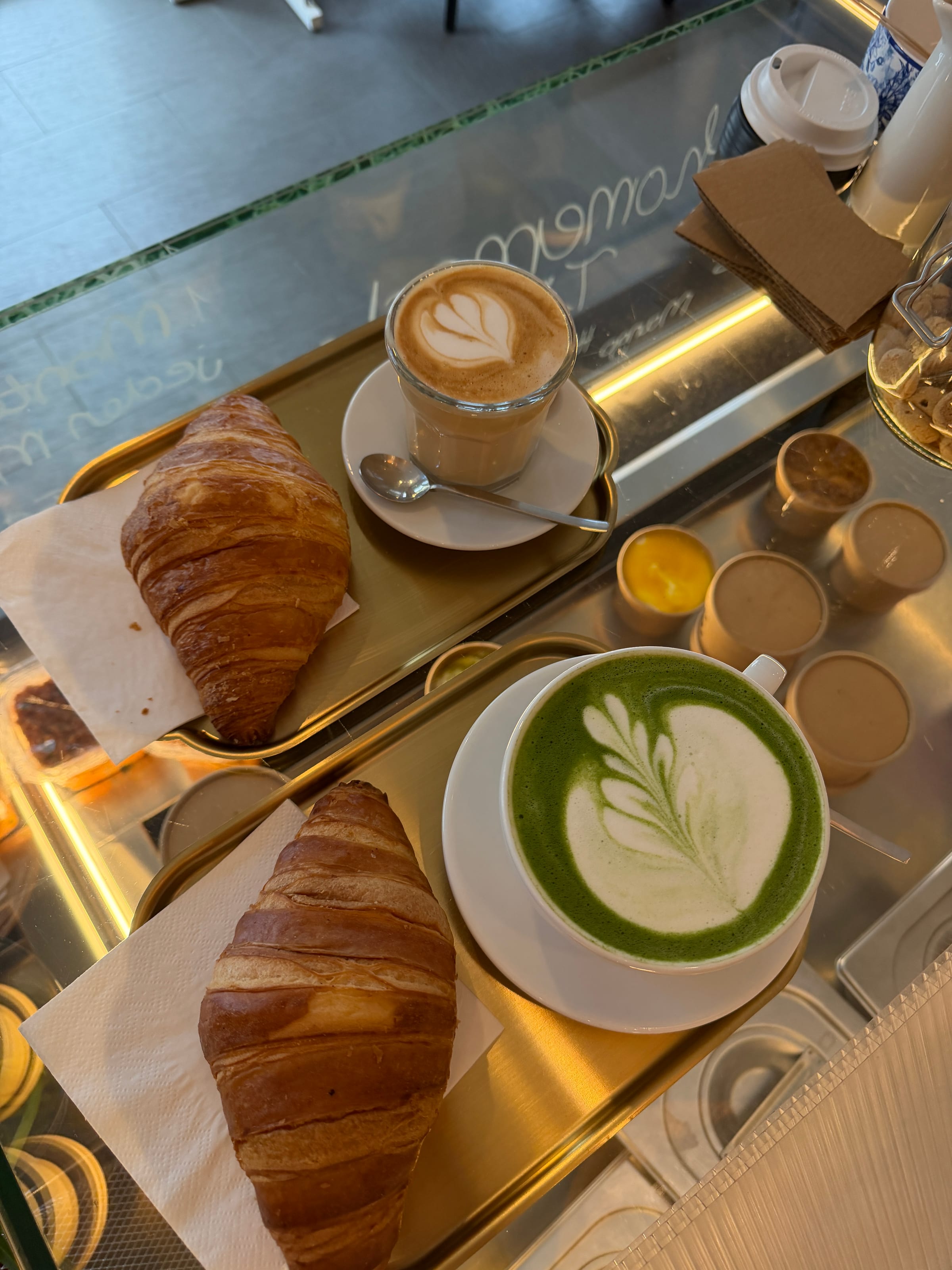 Zwei Buttercroissants, ein Kaffee mit Latte Art im Glas und ein Matcha Latte auf Messingtabletts an der Vitrine